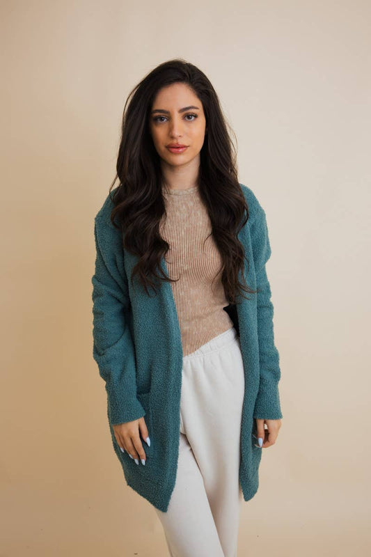 Woman wearing a teal cardigan over a beige sweater and white pants on a beige background