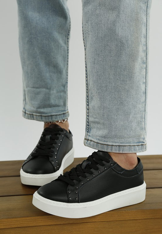 Black sneakers with white soles worn with light blue jeans on a wooden floor.