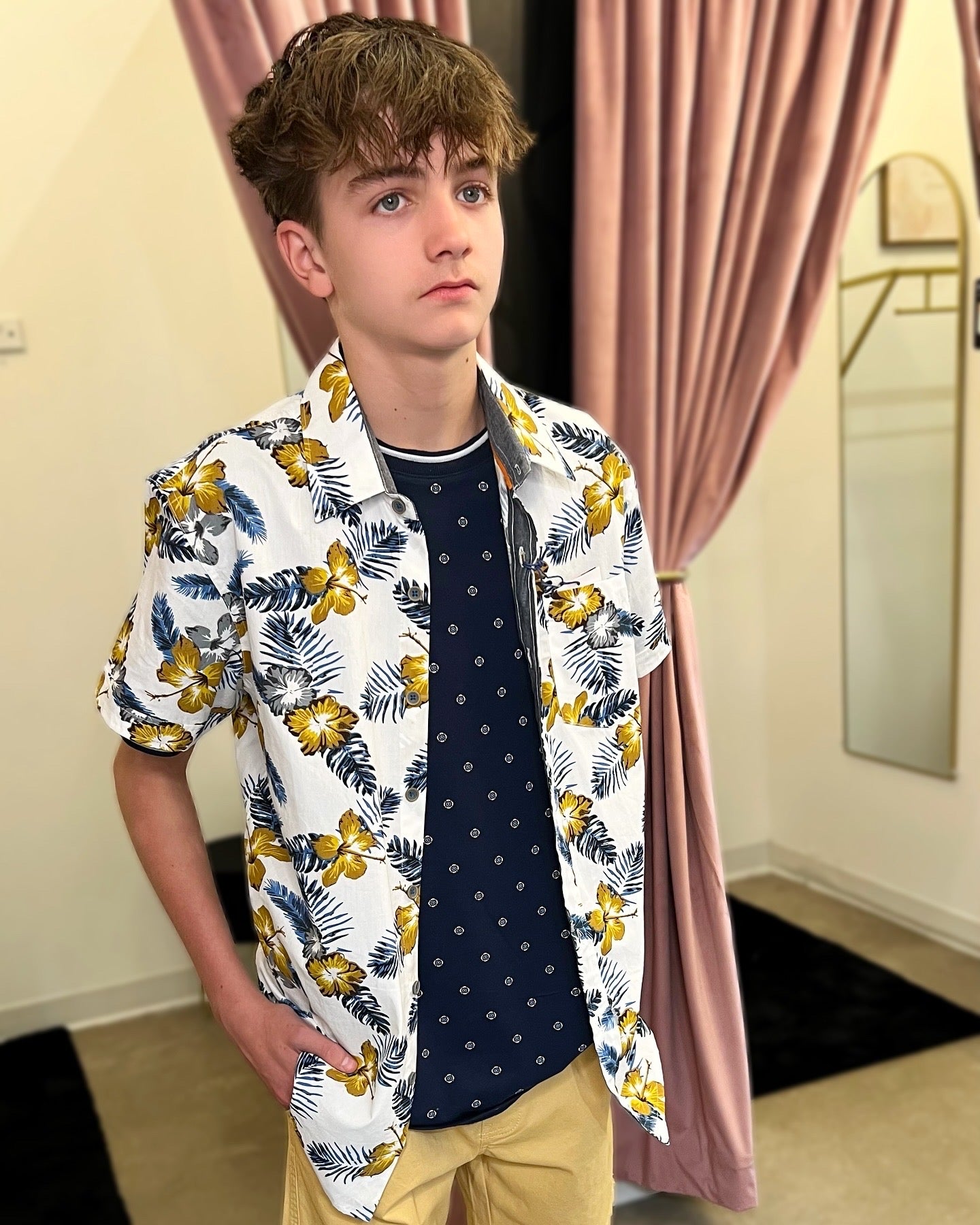 Person wearing a floral shirt over a dark top and yellow pants in a room with pink curtains.