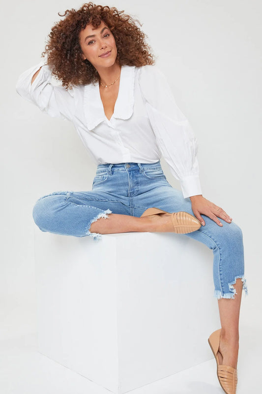 A woman sitting on a bench wearing a white shirt and blue distressed crop jeans with frayed hem, paired with beige strappy sandals.