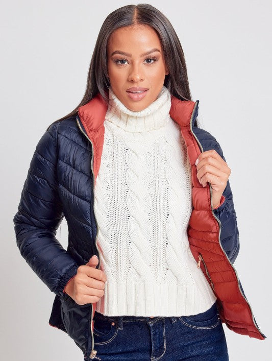 Woman wearing a navy puffer jacket with red lining, white turtleneck sweater, and blue jeans on a light gray background.