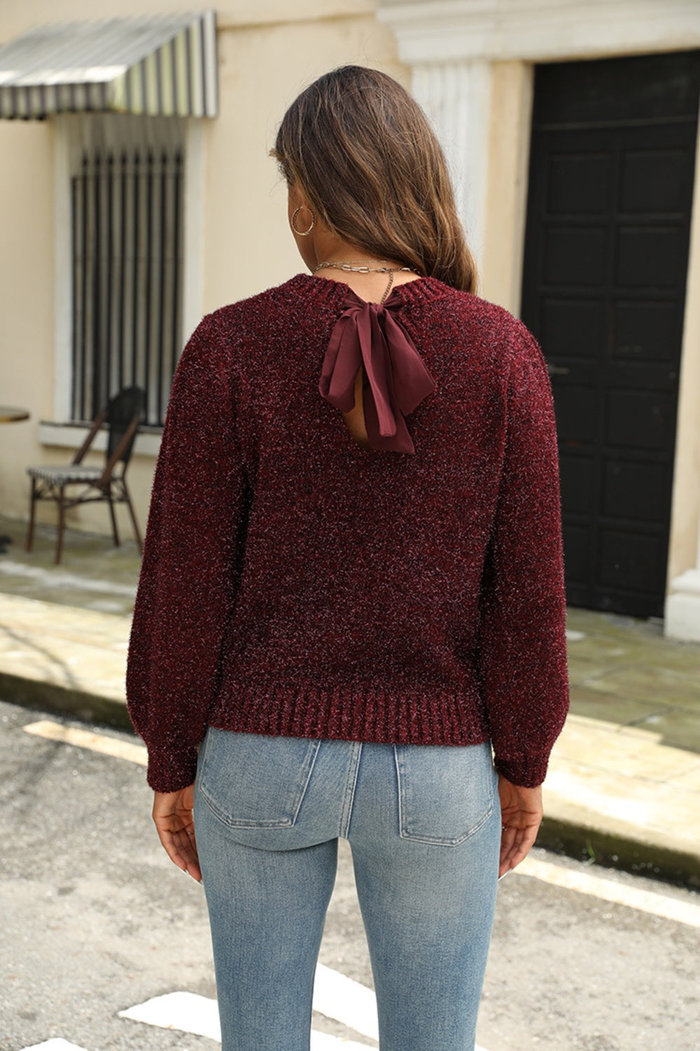 Person wearing a burgundy sweater with a bow detail on the back, standing on a street.
