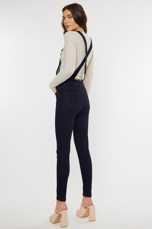 Woman wearing a beige sweater and dark jeans with suspenders on a white background