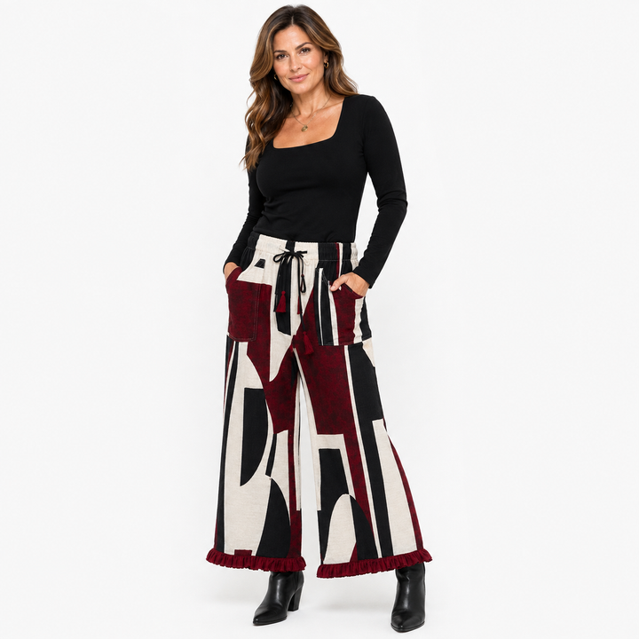 A woman modeling wide-leg pants with an abstract geometric pattern in burgundy, black, and cream, paired with a black long-sleeve top and black ankle boots, against a plain white background.