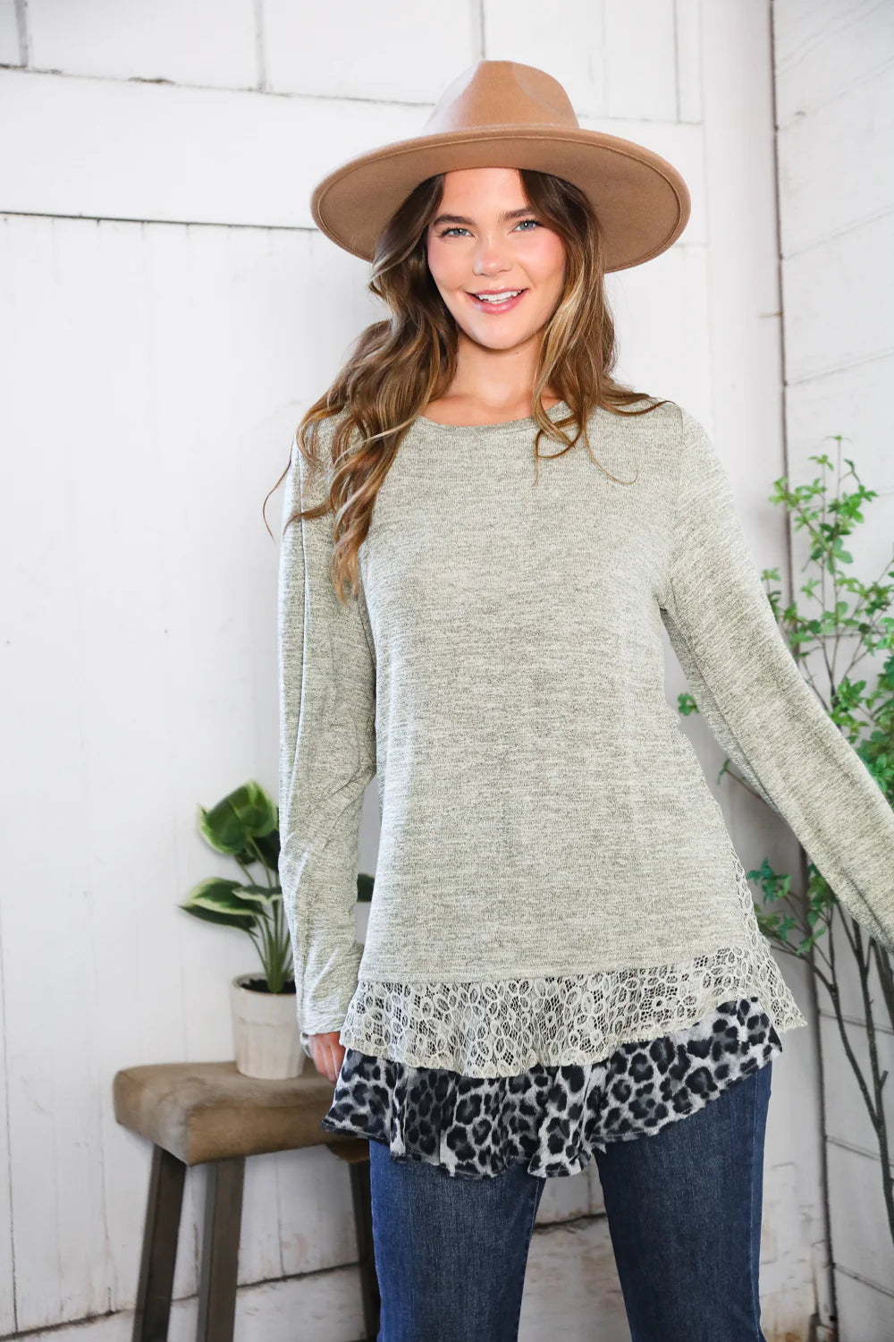 Woman wearing a light green long-sleeve top with a patterned hem, blue jeans, and a brown hat indoors.