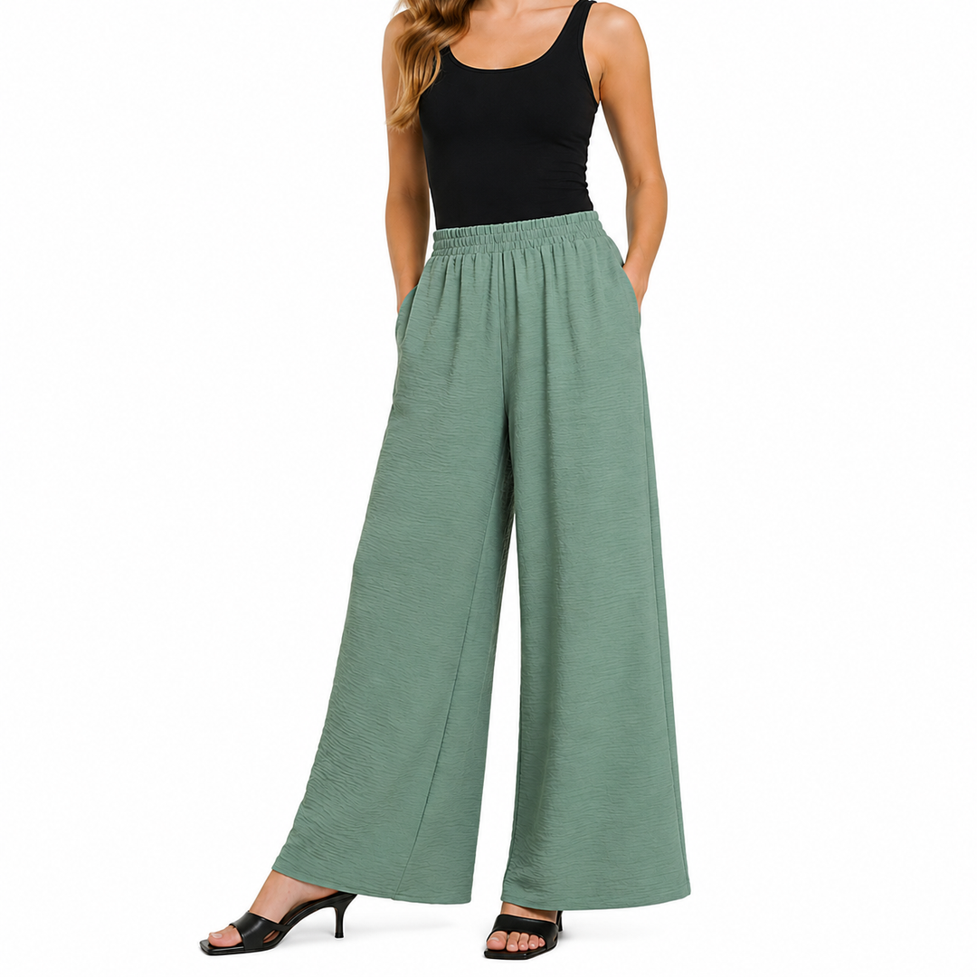 A model wearing a pair of sage green wide-leg pants with an elastic waistband, paired with a black tank top and black heeled sandals, shown against a plain white background.