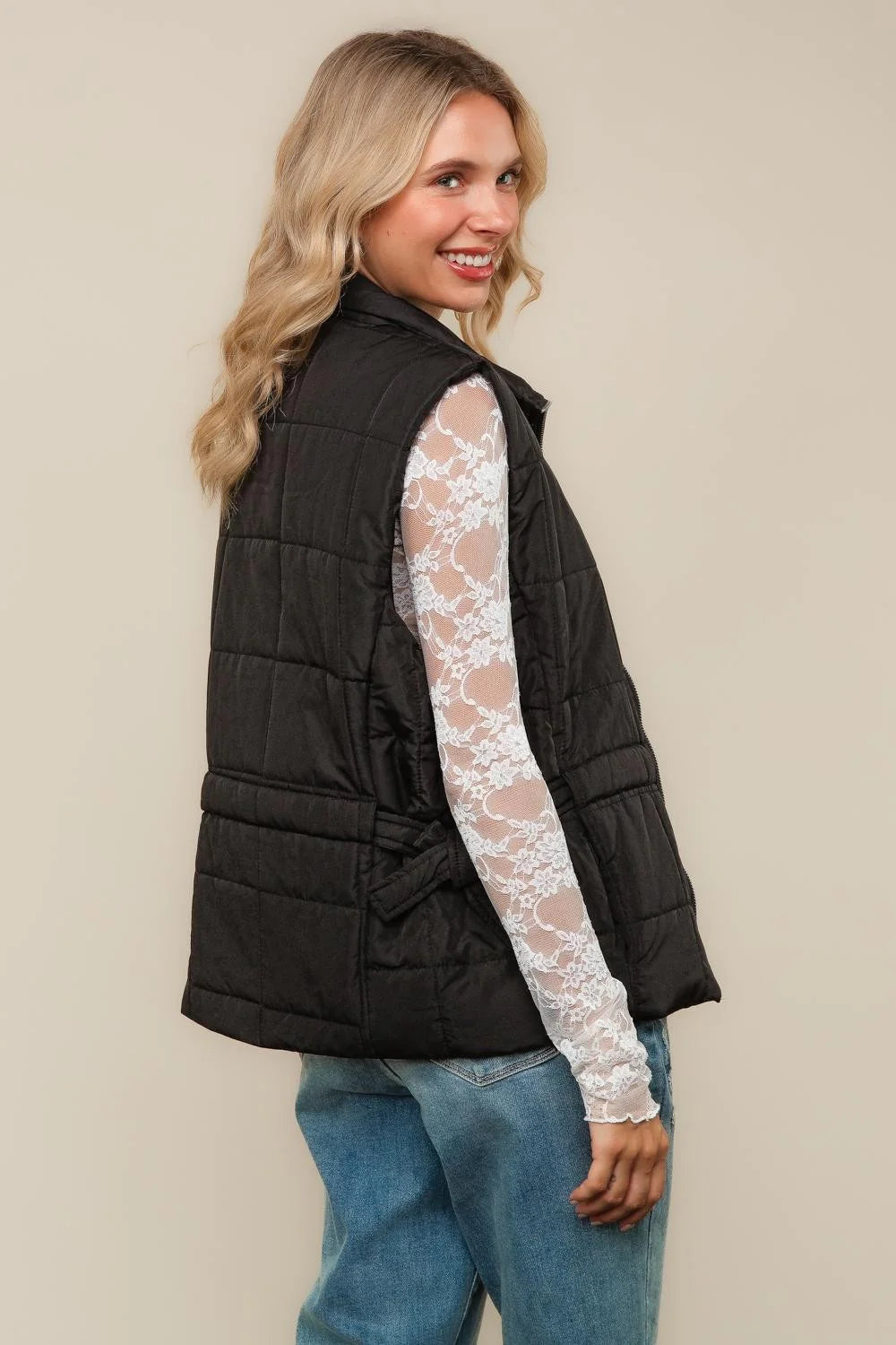 Woman wearing a black puffer vest over a lace long-sleeve top on a beige background