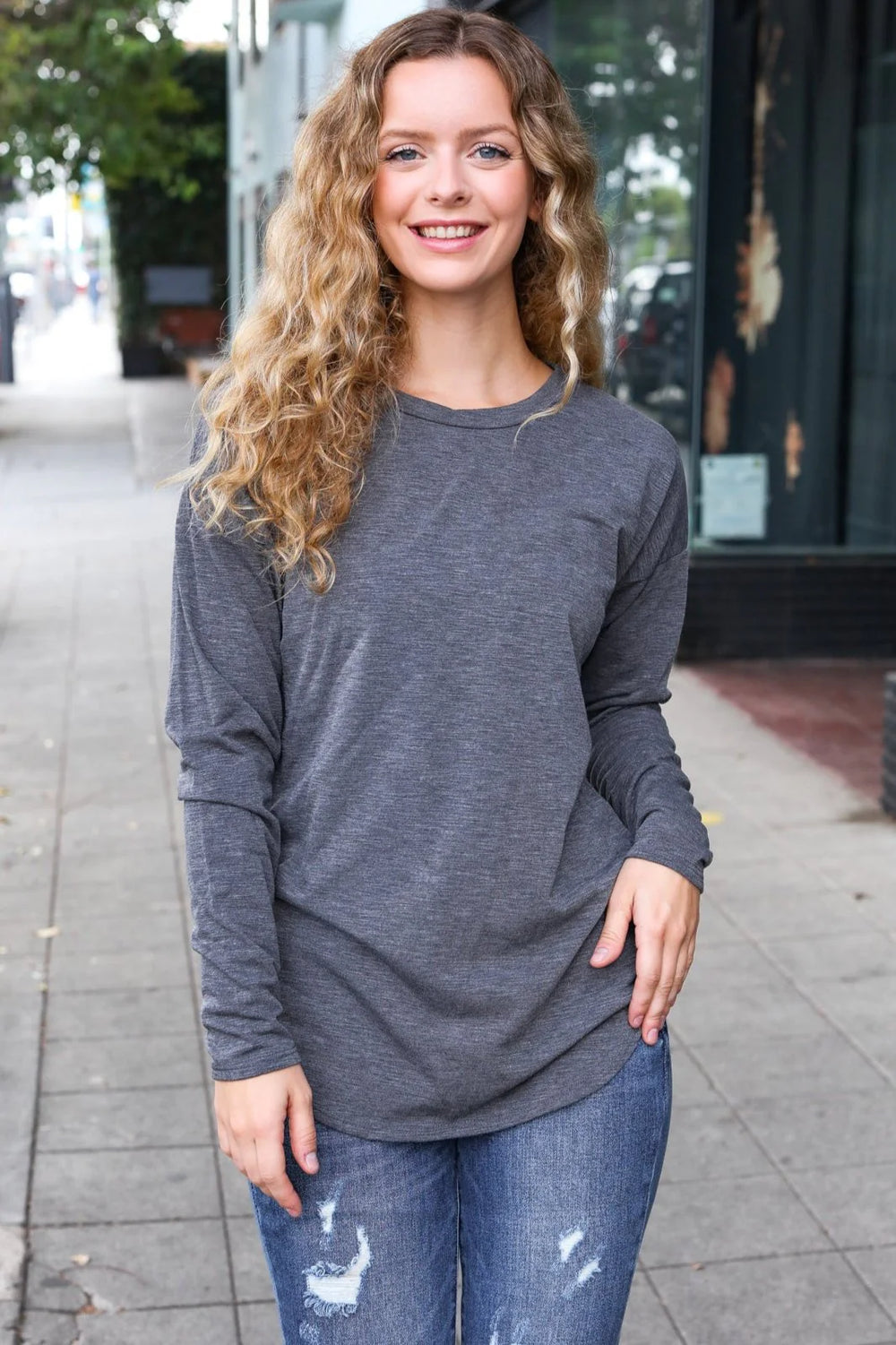 Woman wearing a gray long-sleeve top and blue jeans on a sidewalk.