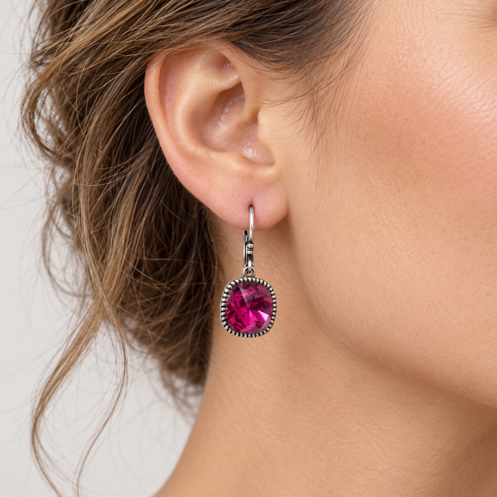 A close-up of a woman's ear wearing a dangling earring with a round bright pink gemstone in a silver-toned bezel setting.