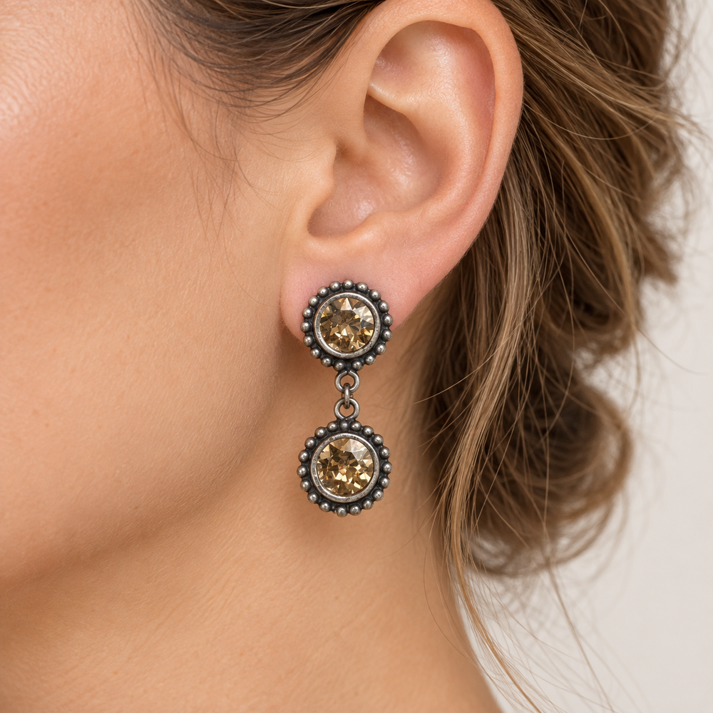 A close-up of a woman's ear wearing a dangling earring with two round amber-colored stones in dark silver-toned bezel settings.