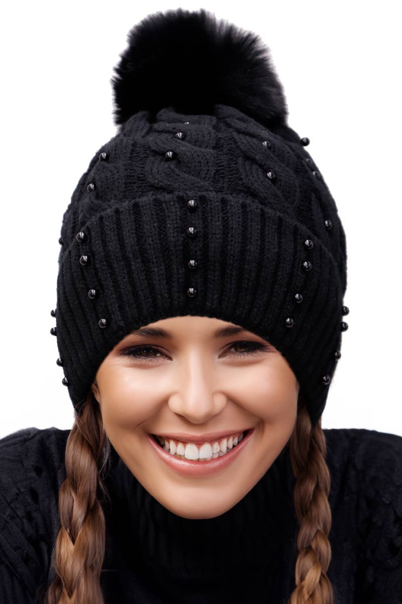 Black knit beanie with pom-pom and embellishments worn by a person on a white background