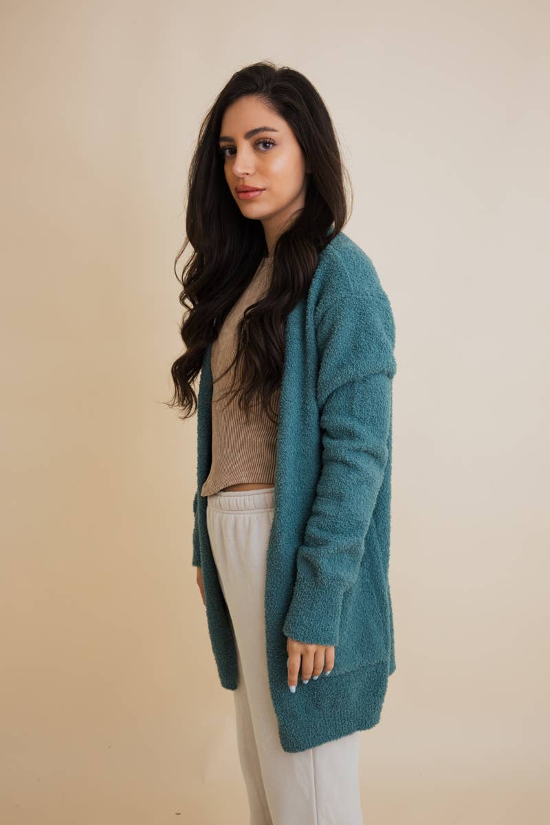 Woman wearing a teal cardigan over a beige sweater with white pants against a beige background