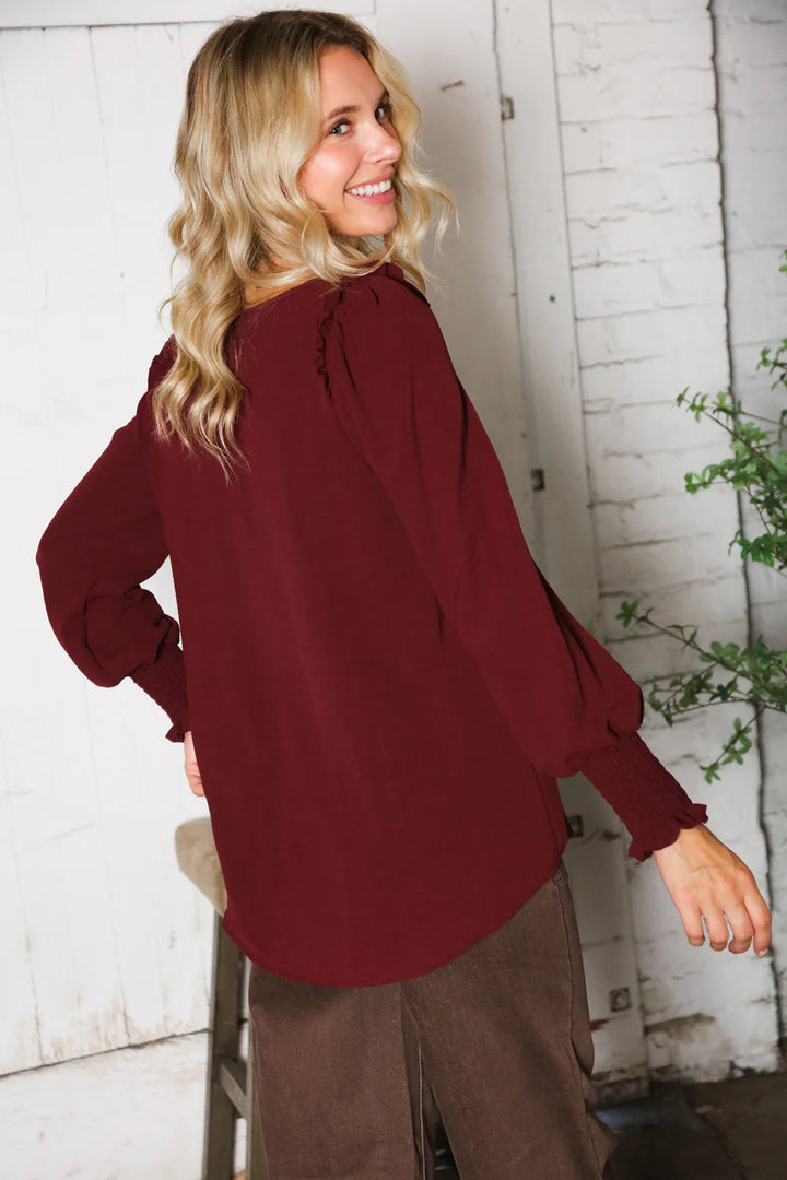 Woman wearing a burgundy blouse and brown pants standing against a white brick wall.