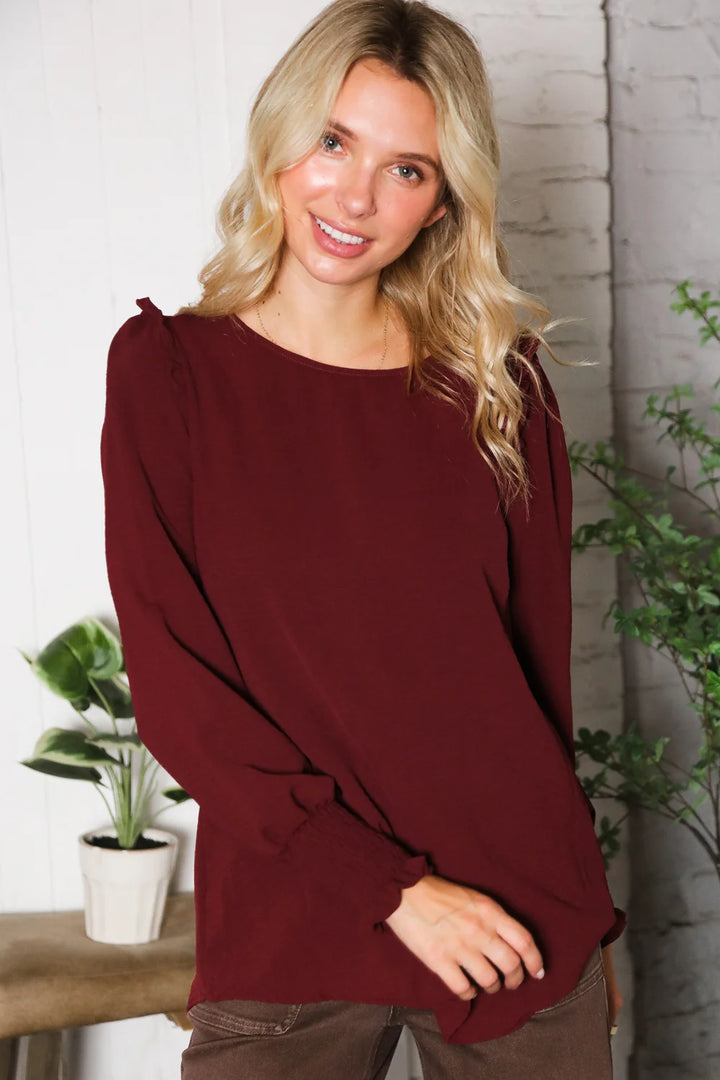 Woman wearing a burgundy top with a white brick wall and plants in the background
