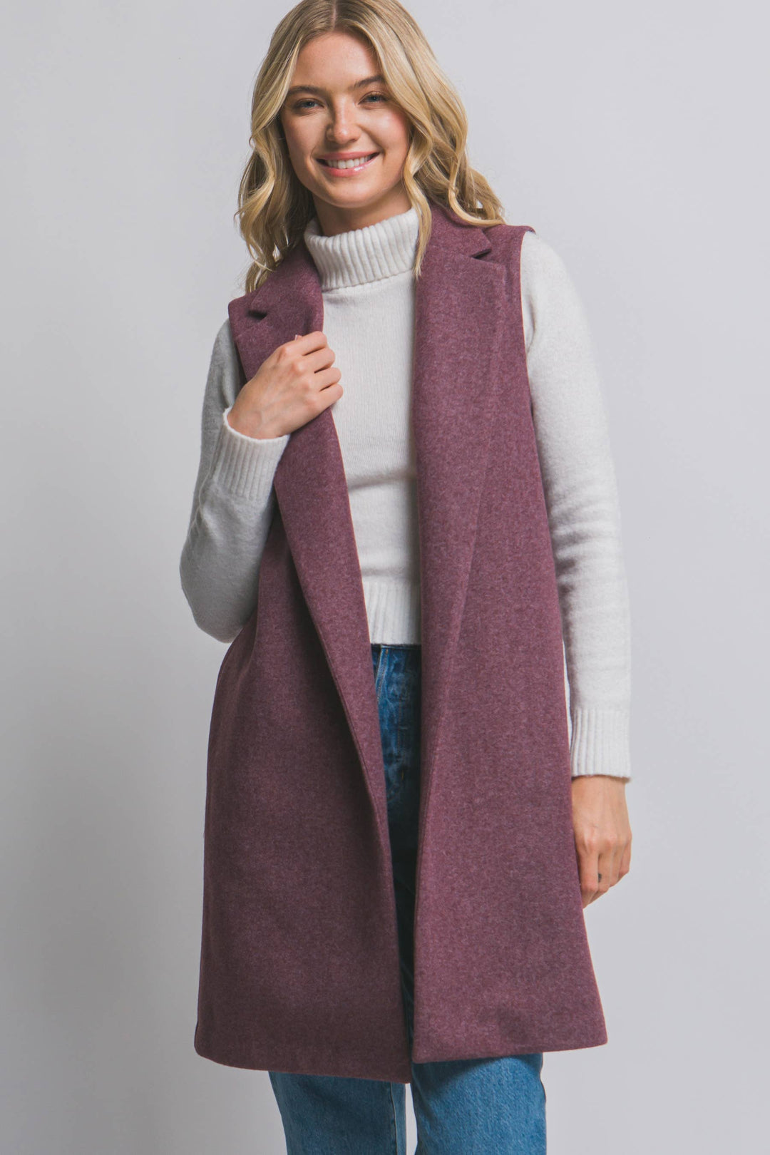 Woman wearing a long purple vest over a white sweater and blue jeans on a plain background