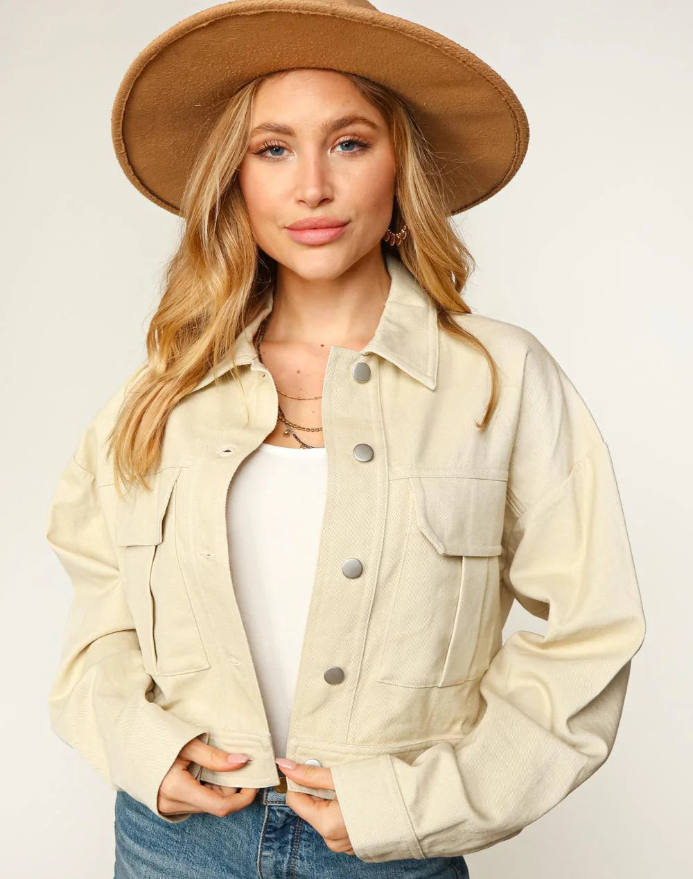 Woman wearing a beige jacket, white top, and denim shorts with a brown hat on a plain background