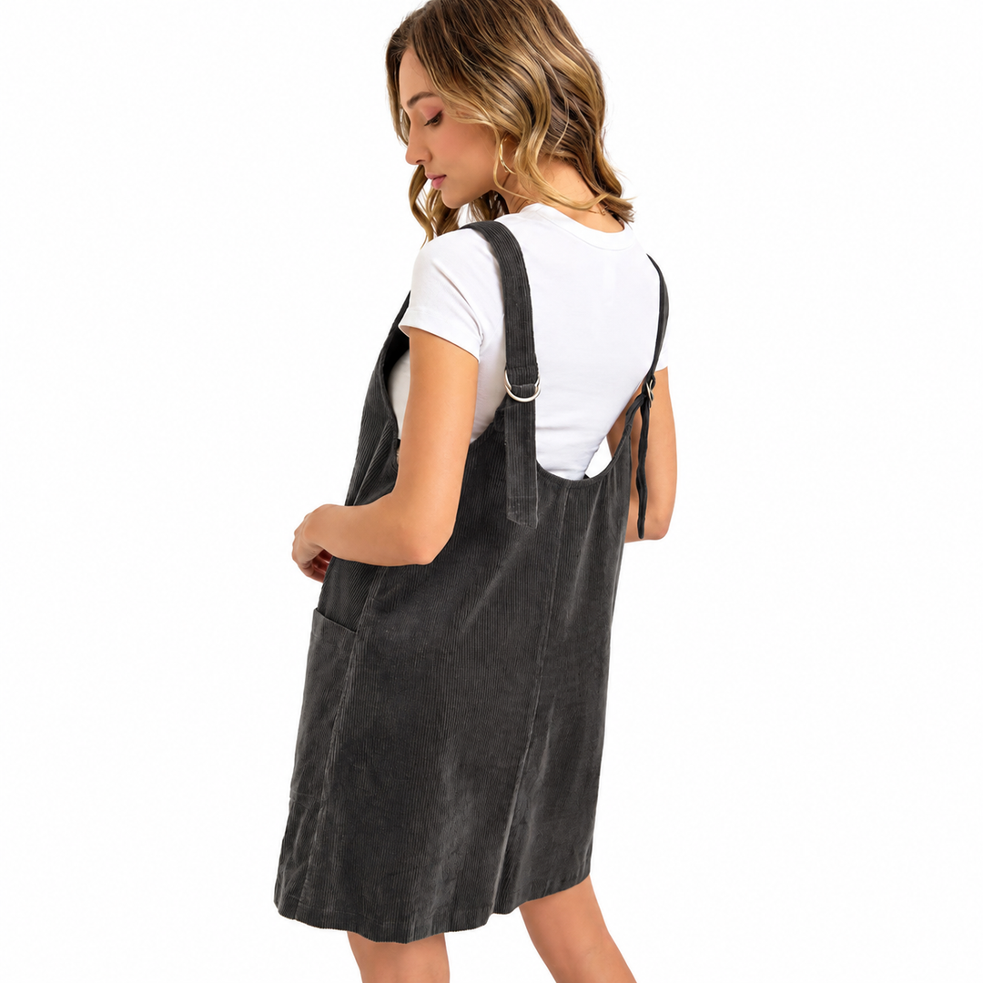 A woman wearing a dark gray corduroy pinafore dress over a white t-shirt, shown from a three-quarters back view against a solid white background.