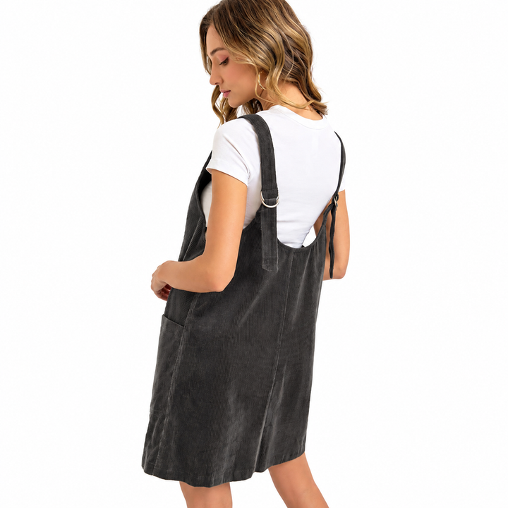 A woman wearing a dark gray corduroy pinafore dress over a white t-shirt, shown from a three-quarters back view against a solid white background.