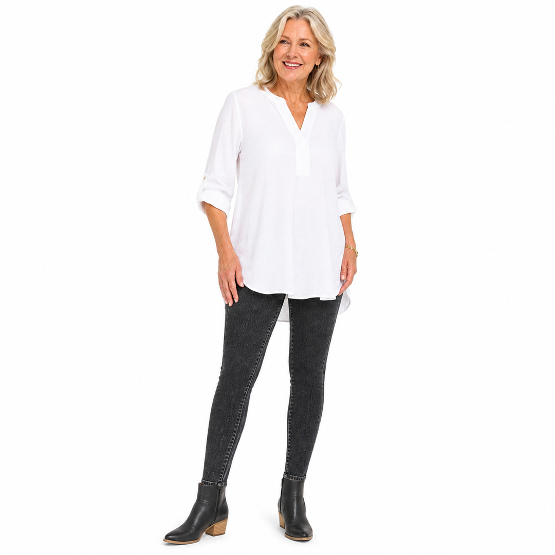 A smiling blonde woman stands against a plain white studio background wearing a loose white long-sleeve blouse with rolled cuffs, dark gray skinny jeans, and black ankle boots.
