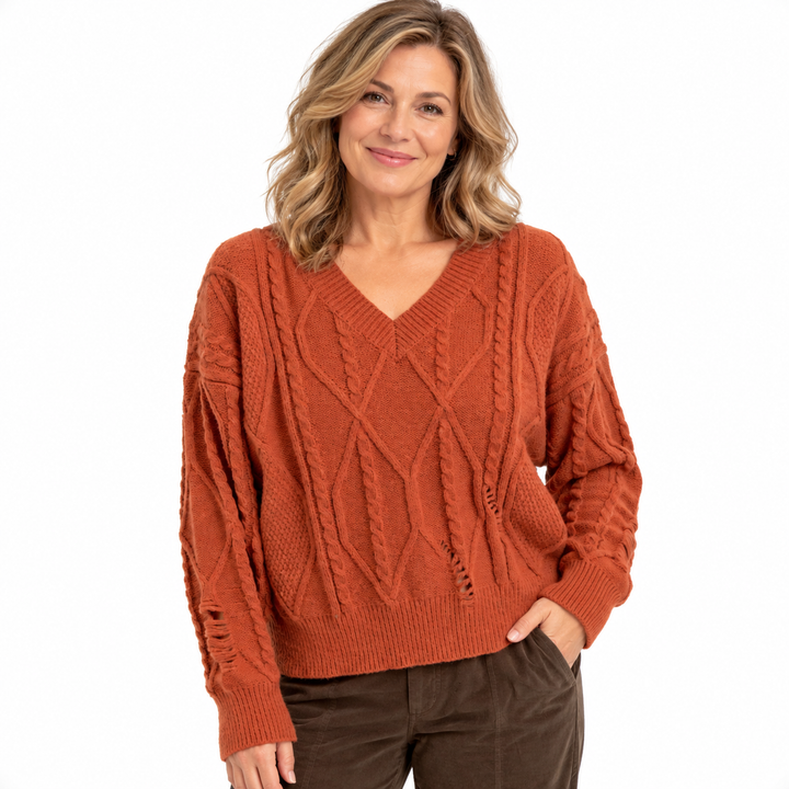 A woman wearing a rust-orange cable-knit V-neck sweater with distressed details on the sleeves, paired with dark brown pants, against a plain white background.