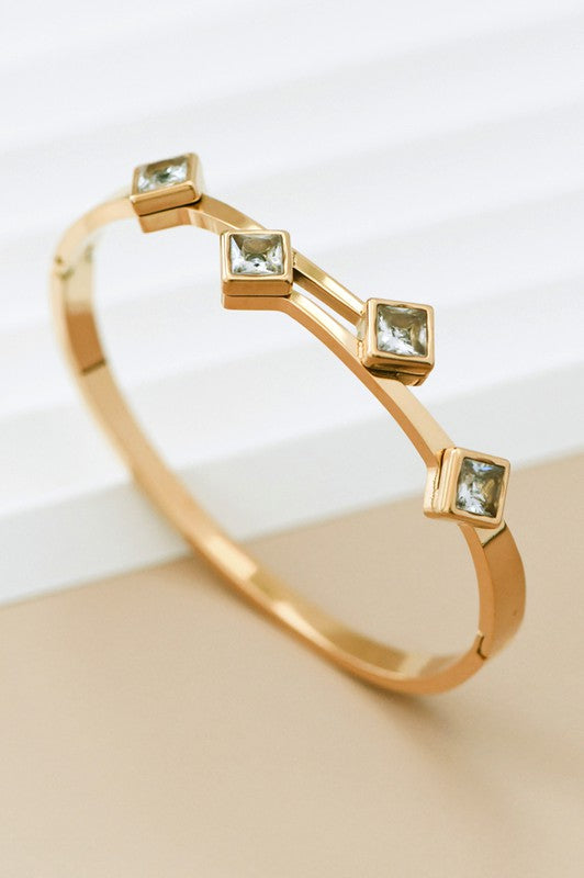 A gold hinged bracelet with multiple square stone accents on a light surface.