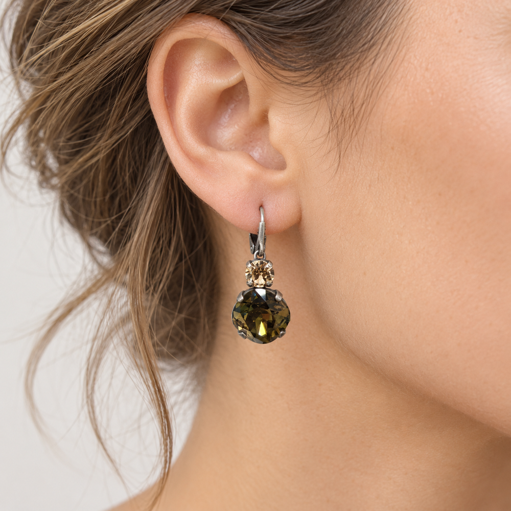 A close-up of a woman's ear wearing a dangling earring with a faceted olive green gemstone and a small clear accent stone in a silver-toned setting.