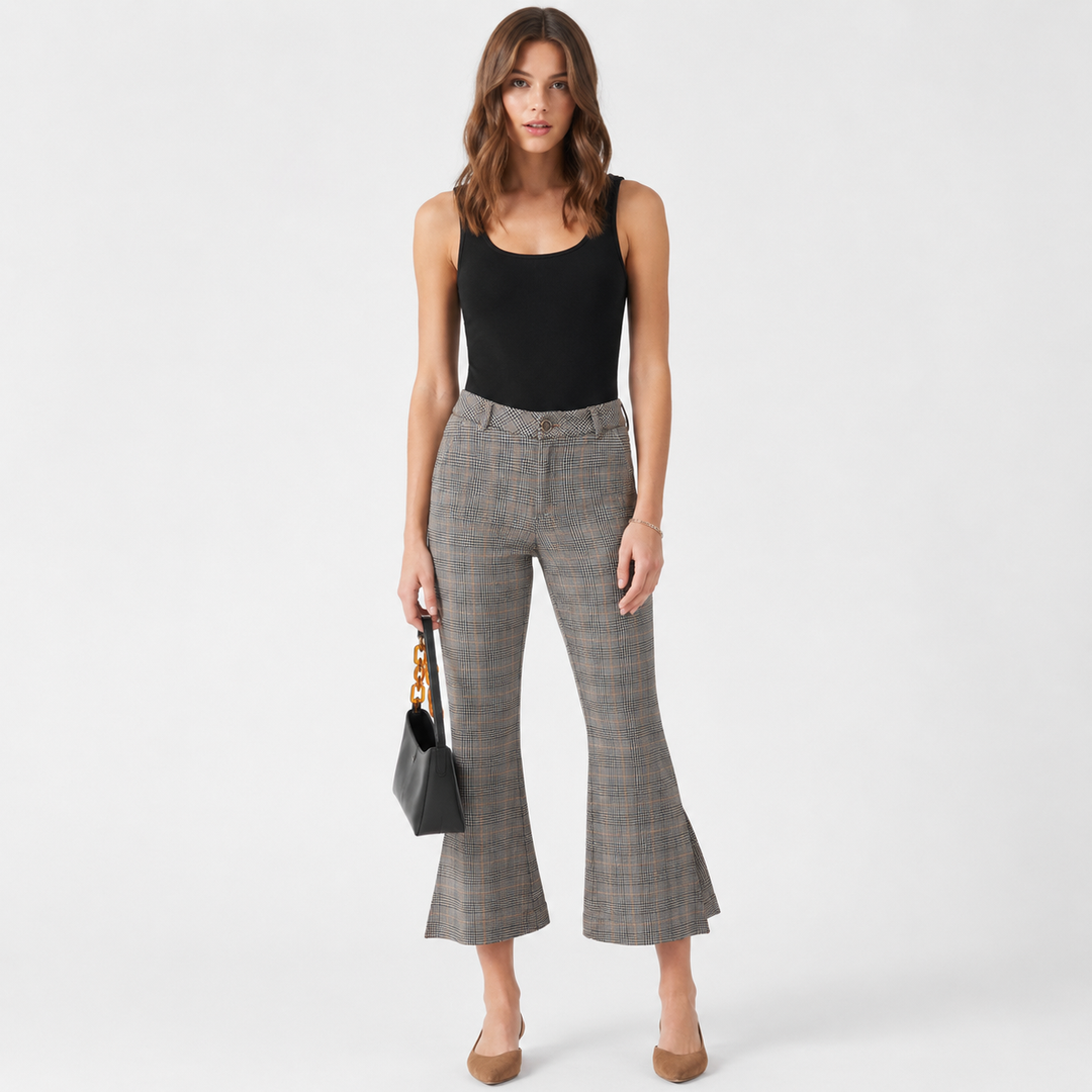 A woman modeling a pair of gray and black plaid cropped flare pants with a black sleeveless top, holding a small black handbag against a light gray studio background.