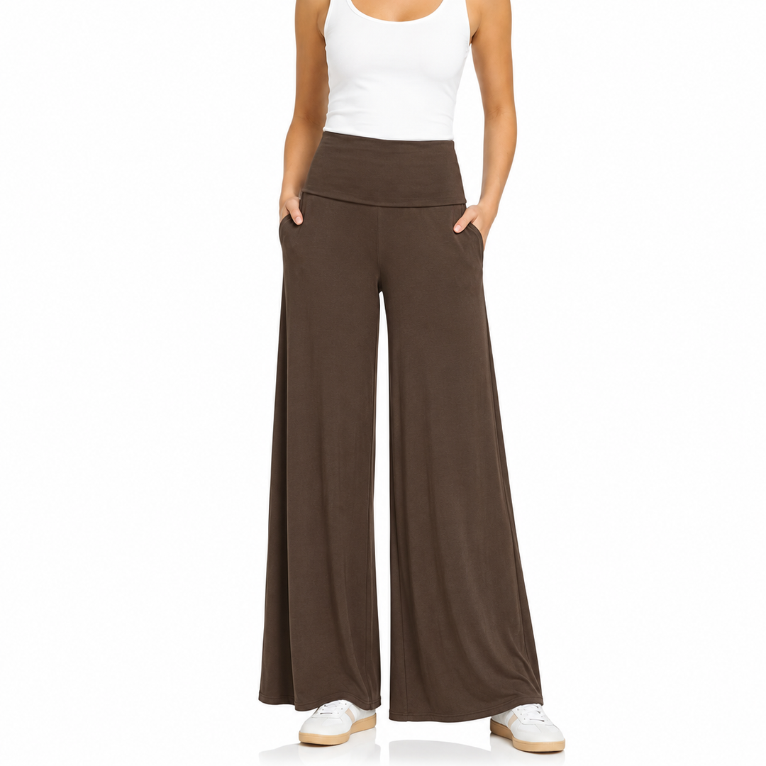 A model wearing high-waisted, wide-leg brown pants with a matching elastic waistband, styled with a white tank top and white sneakers against a plain white background.