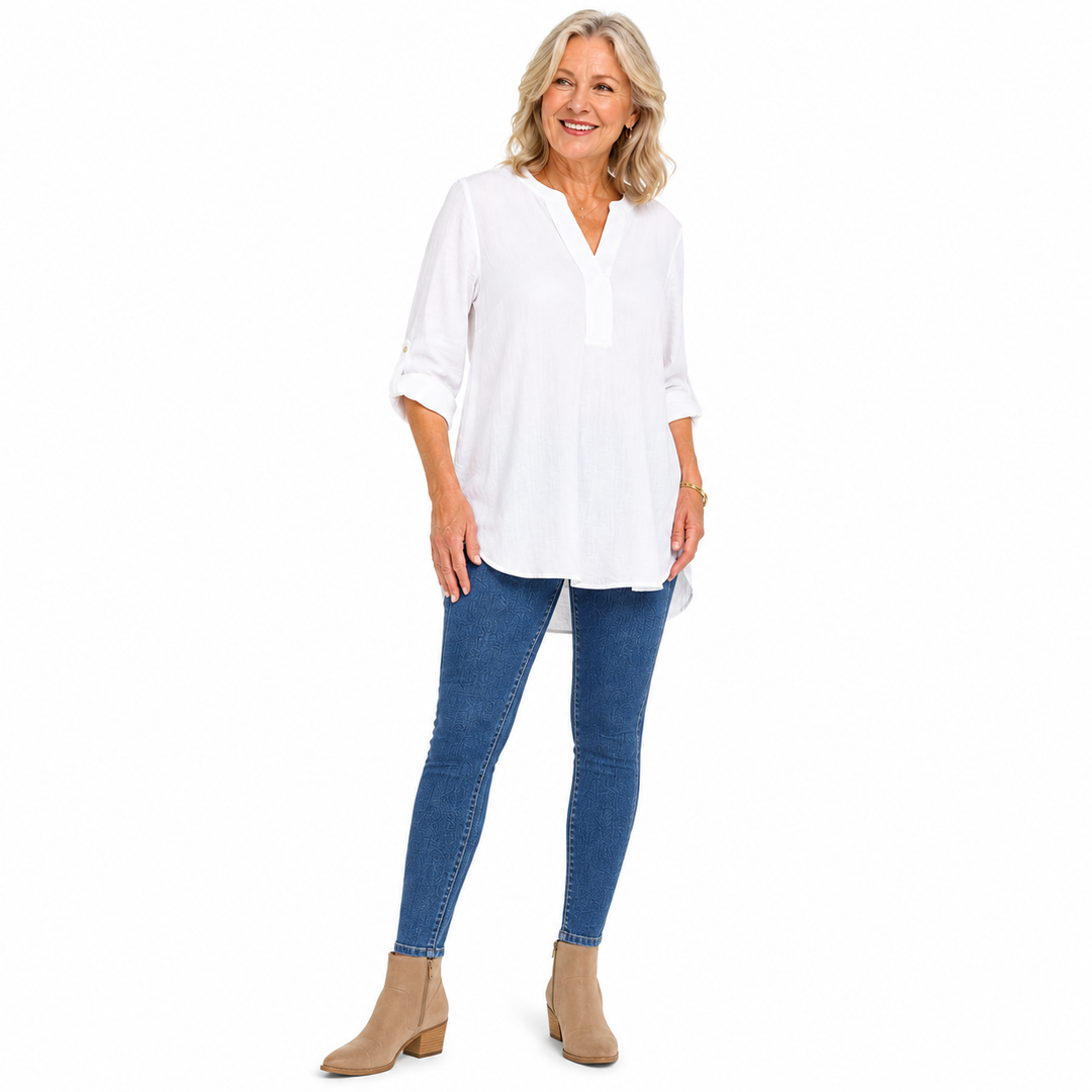 A smiling woman stands against a plain white studio background wearing a loose white V-neck blouse with rolled sleeves, blue skinny jeans, and tan ankle boots.