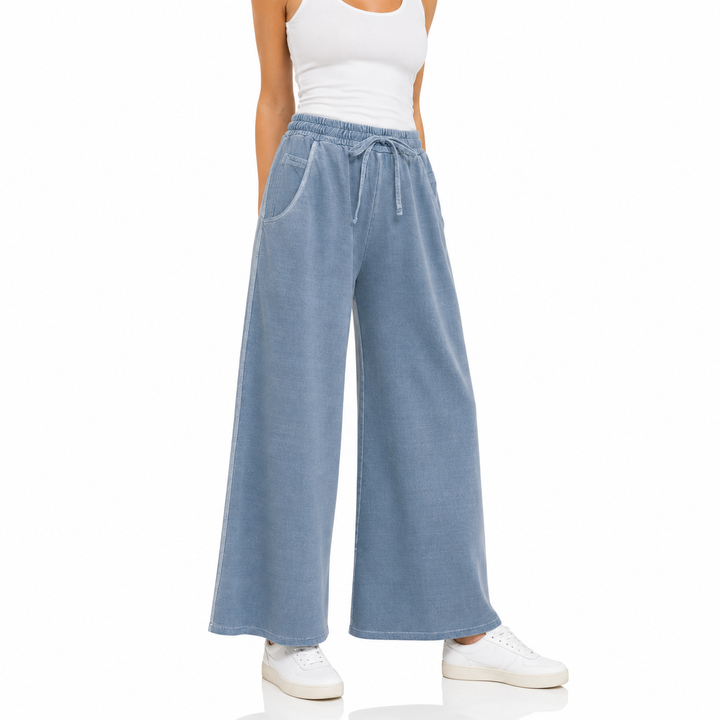 A model wearing light blue wide-leg denim pants with a drawstring waist, paired with a white tank top and white sneakers, shown against a plain white background.