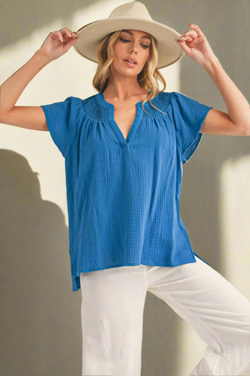 Woman wearing a blue top and white pants with a beige hat against a plain background