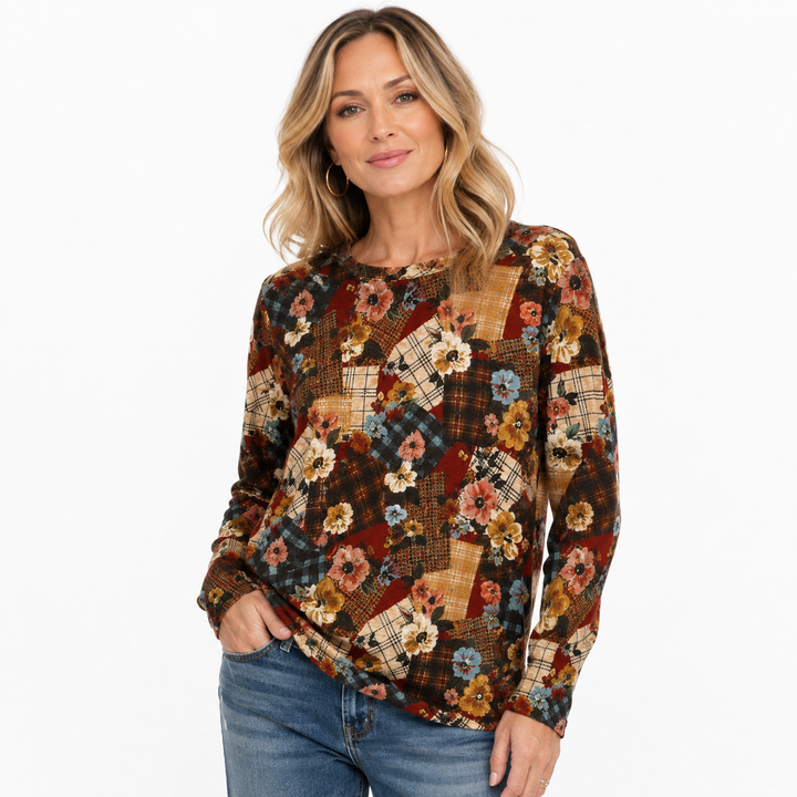 A woman modeling a long-sleeve patchwork top with a floral and plaid pattern in earthy tones, paired with blue jeans, against a plain white background.