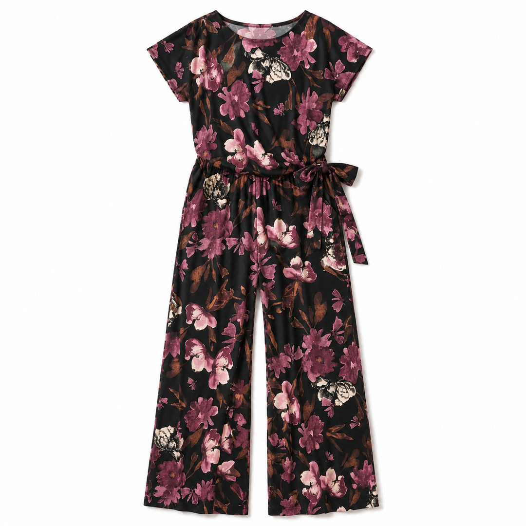 A short-sleeve jumpsuit with a wide-leg pant design, featuring a dark brown base covered in a pink, purple, and white floral print. It has a matching fabric tie belt at the waist and is displayed flat against a solid white background.
