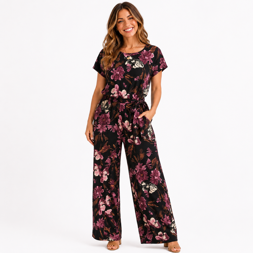 A woman modeling a short-sleeve jumpsuit with a dark floral print featuring pink, purple, and white flowers on a black background, set against a plain white studio backdrop.