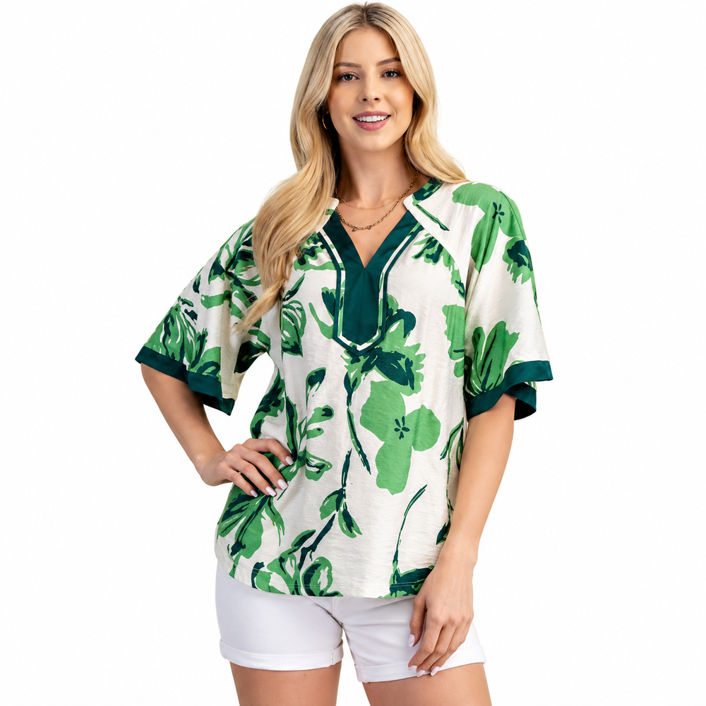 A woman modeling a short-sleeve V-neck top with a green tropical floral print on an off-white background, paired with white shorts, against a plain white studio background.
