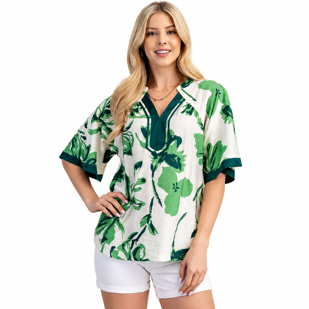 A woman modeling a short-sleeve V-neck top with a green tropical floral print on an off-white background, paired with white shorts, against a plain white studio background.