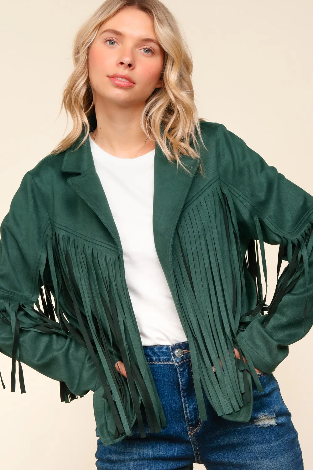 Woman wearing a green fringe jacket over a white shirt and blue jeans on a beige background