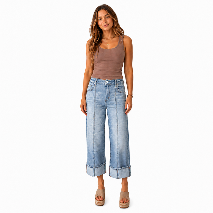 A woman modeling a pair of light-wash wide-leg cropped jeans with rolled hems, paired with a brown tank top and beige platform sandals, against a plain white background.