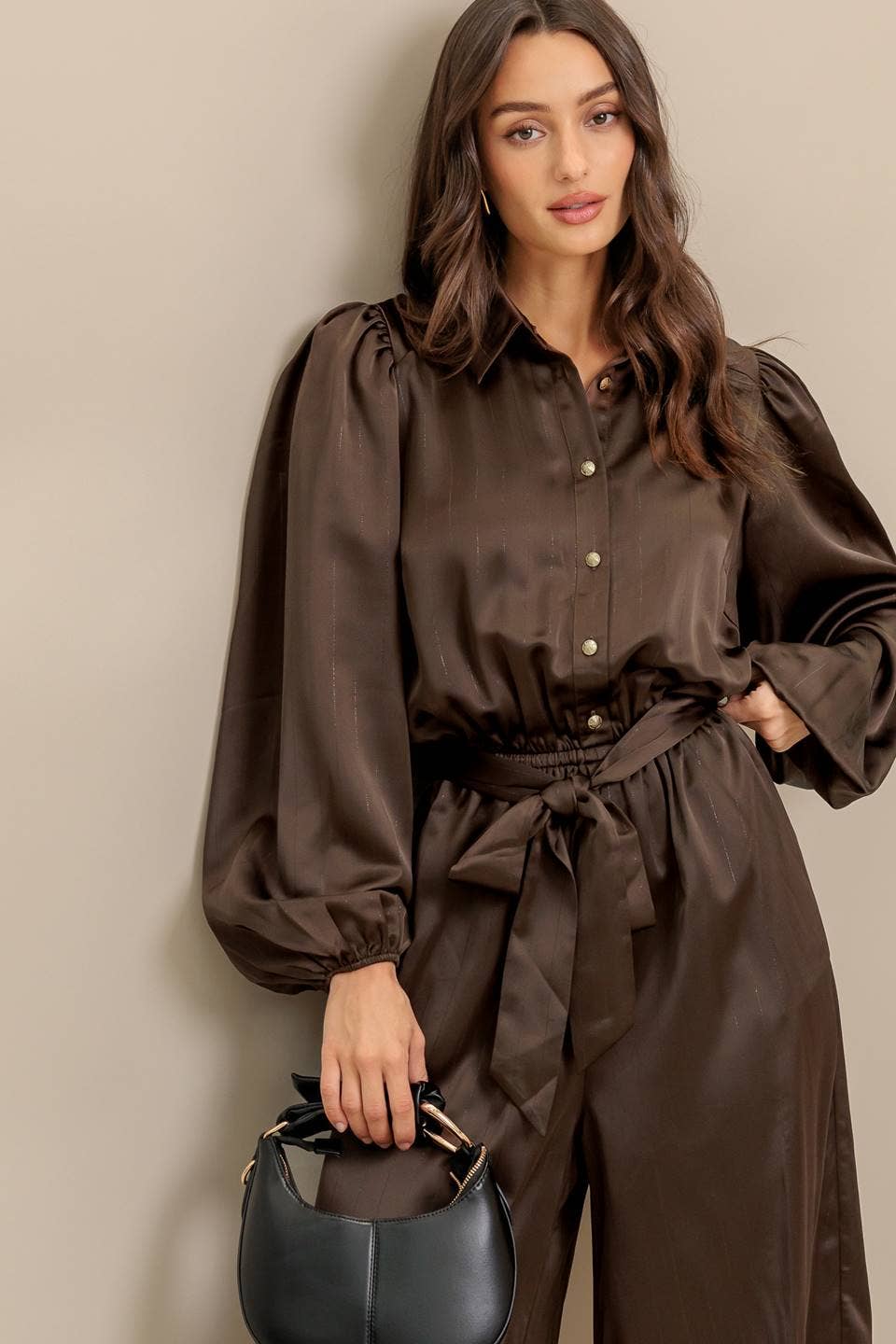 Woman wearing a brown jumpsuit holding a black handbag against a beige wall