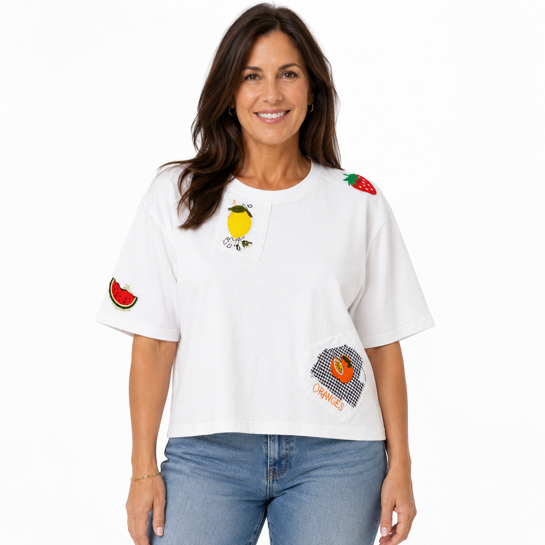 A woman wearing a white short-sleeve crop top T-shirt with colorful fruit graphics (lemon, strawberry, watermelon, orange) and blue jeans, photographed against a solid white background.