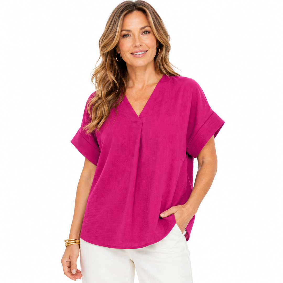 A woman modeling a bright magenta short-sleeve V-neck top with a gathered front detail, paired with white pants, against a plain white background.