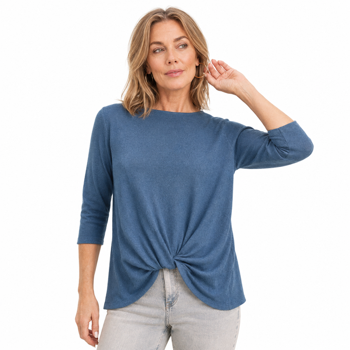 A woman modeling a slate blue three-quarter sleeve top with a twisted knot detail at the hem, paired with light gray jeans, against a solid white background.