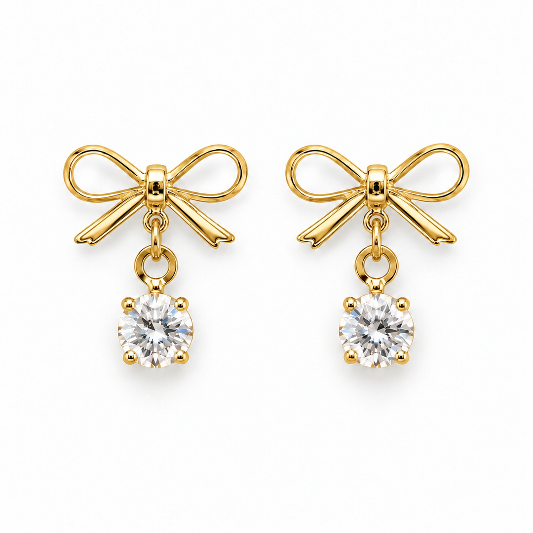 A pair of gold bow stud earrings with a clear round-cut gemstone dangling below each bow, shown on a plain white background.