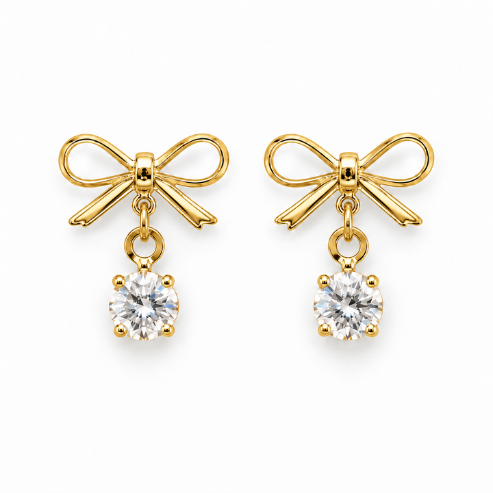 A pair of gold bow stud earrings with a clear round-cut gemstone dangling below each bow, shown on a plain white background.