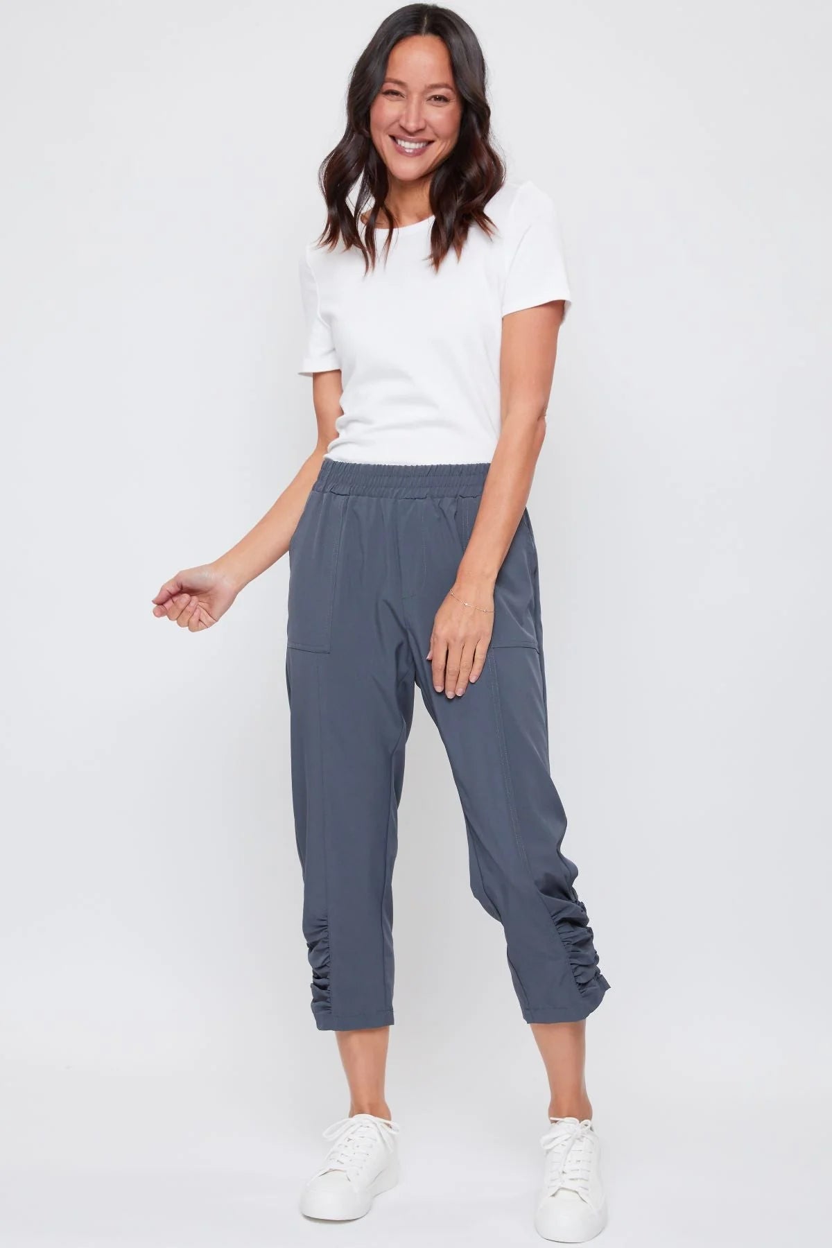 A woman is standing, wearing capri-length pants with a side rouching detail and an elastic waistband, paired with a black tank top and white sneakers.