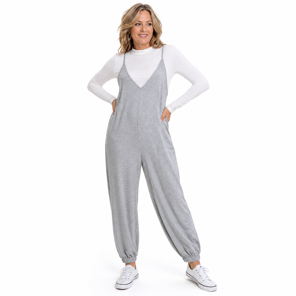 A woman modeling a light gray heathered sleeveless jumpsuit over a white long-sleeve t-shirt and white sneakers, standing against a solid white background.