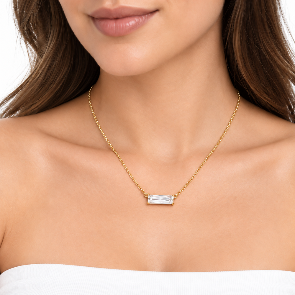 A close-up of a woman wearing a delicate gold chain necklace with a small rectangular clear gemstone pendant, shown against her neck and upper chest.
