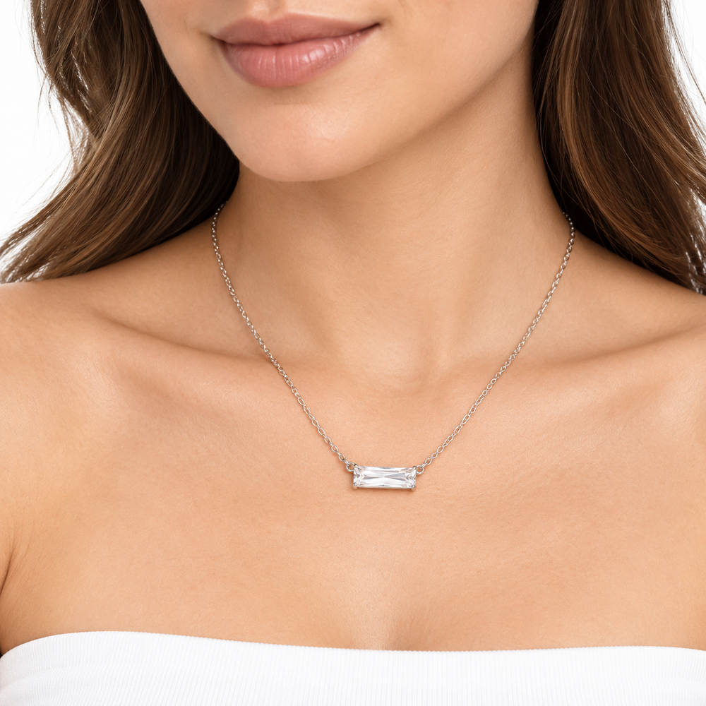 A close-up of a woman wearing a delicate silver chain necklace with a small rectangular clear gemstone pendant, shown against her neck and upper chest.
