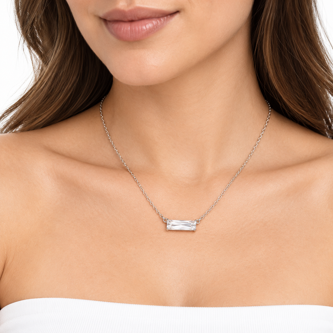 A close-up of a woman wearing a delicate silver chain necklace with a small rectangular clear gemstone pendant, shown against her neck and upper chest.