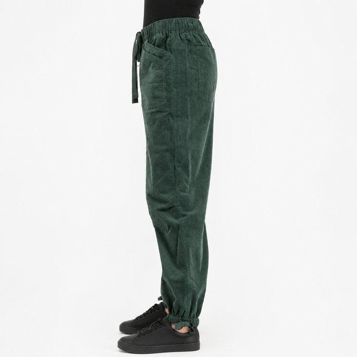 A side view of a person wearing dark green corduroy jogger pants with an elastic waist and drawstring, paired with black sneakers, against a plain white background.